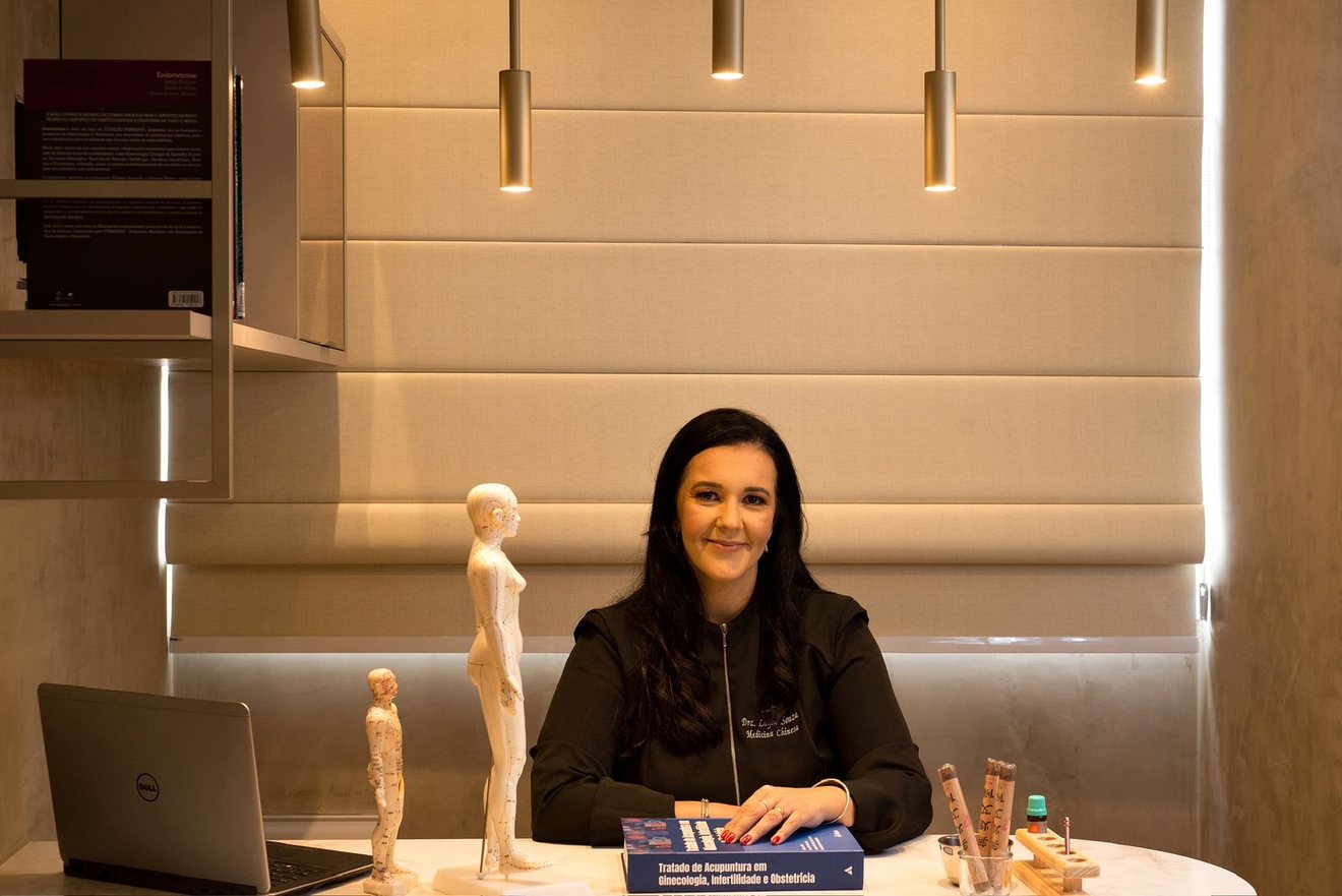 Layla Souza à mesa do consultório, com modelos de acupuntura e um tratado de acupuntura em ginecologia